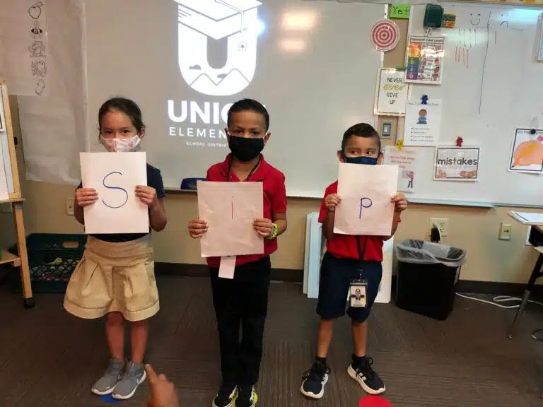 Kindergarten students holding letters “s,” “i,” and “p” written on white paper. The “i” is written in a different color.